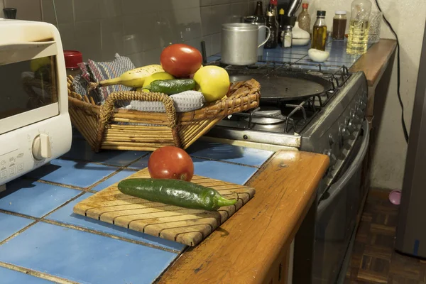 Birçok taze sebze mutfaktaki ahşap bir kesme tahtasının üzerinde oturur. Sepette daha fazla sebze var. Arka planda bir soba ve diğer mutfak malzemeleri var..