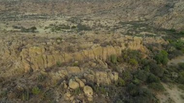 Görüntüler, gün batımında bir dağın yakınındaki kuru bir yerde kaktüslü kayalık bir alan gösteriyor. Manzara engebelidir ve yerde doğal oluşumlar gösterir..