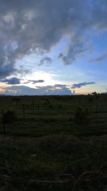 Güneş akşam geniş yeşil bir tarlanın üzerinde batar. Bulutlar gökyüzünü doldurarak, ışık ve gölgenin bir karışımını oluşturuyor. Manzara bitkilerin ve uzak tepelerin sıralarını gösteriyor..