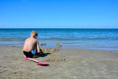 Arkadan küçük sarışın çocuk, yüzme şortuyla kumsalda kumdan bir kale inşa ediyor, denizin önünde, mavi gökyüzü ile.