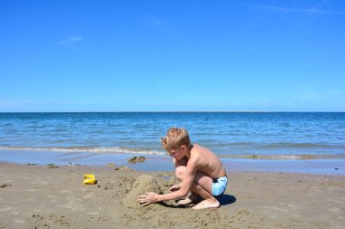 Yüzme şortlu sarışın bir çocuk kumsalda kumdan bir kale inşa ediyor. Denizin önünde, mavi gökyüzü olan.