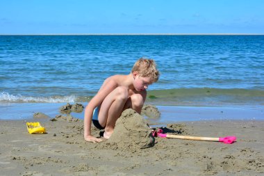 Yüzme şortlu sarışın bir çocuk kumsalda kumdan bir kale inşa ediyor. Denizin önünde, mavi gökyüzü olan.
