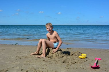 Yüzme şortlu sarışın bir çocuk kumsaldaki kumların üzerinde oturmuş gülüyor, mavi denizin önünde.