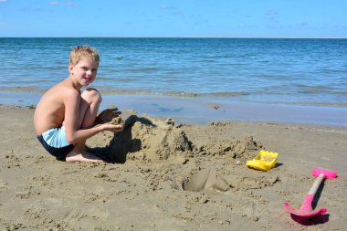 Yüzme şortlu küçük sarışın çocuk kumsalda kumdan bir kale inşa ediyor denizin önünde, mavi gökyüzü ile