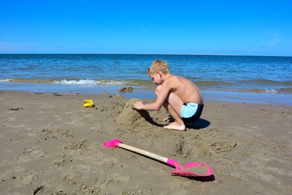 Blondyn Kąpielówkami Buduje Zamek Piasku Plaży Przed Morzem Błękitnym Niebem — Zdjęcie stockowe
