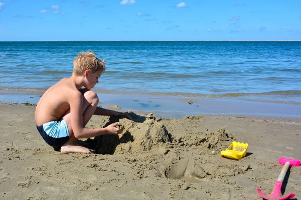 Blondyn Kąpielówkami Buduje Zamek Piasku Plaży Przed Morzem Błękitnym Niebem — Zdjęcie stockowe