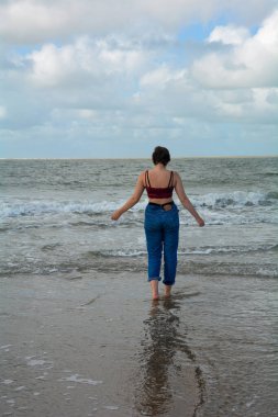 Kot pantolonlu ve kısa bluzlu genç kız kumlu sahilde yalınayak koşuyor mavi gökyüzü ve bulutlarla denize doğru.