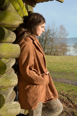 Yandan genç bir kadın, açık kahverengi ceketli, ahşap bir kulübeye yaslanmış, başı aşağıda, yüzünde ciddi bir ifadeyle ve gözleri kapalı, güneşin tadını çıkarıyor, yeşil doğada.