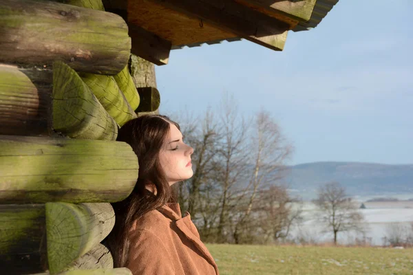 Young woman from the side, with light brown coat, leaning against a wooden hut, with a serious expression on her face and closed eyes, enjoying the sun, in green nature