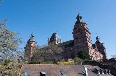 Aschaffenburg, Almanya 'daki Johannisburg Kalesi' nin manzarası mavi gökyüzü ve ön planda eski bir evin çatısı var..