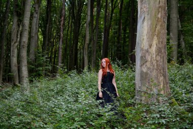 Uzun siyah elbiseli genç kızıl saçlı bir kadın yeşil ormanın ortasında büyük bir ağacın yanında duruyor..