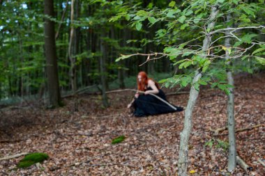 Ön planda bir dal, arka planda, odak dışı, uzun siyah elbiseli, oturan kızıl saçlı bir kadın, ormanda bir yay ve ok ile.