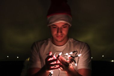 Genç adam gerçek bir Avrupa Noel atmosferinin tadını çıkarıyor ve bir Noel ağacına taktığı küçük ışıklara gülümsüyor. Rahatlatıcı ve huzurlu. Mutlu noeller. Beyaz tişörtlü adam.