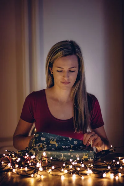 girl of European ethnicity with blond hair with interest unwraps a gift for Christmas or birthday in the warm light of small lights. Holidays, well-being and cohesion.