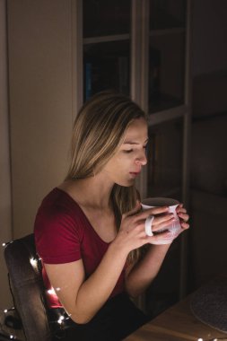 Saçları röfleli bir kadın çayı kabul edilebilir sıcaklıkta soğutmak için fincana üflüyor. Kahverengi gözlü ve güzel bordo bir tişörtü olan çekici bir genç kız. Noel ışıkları.
