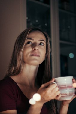 Masada oturan kahverengi gözlü sihirli bir sarışının portresi. Kız, donmuş ellerini bir kupa sıcak çayla ısıtarak pencereden dışarı bakıyor. Koronavirüsle birlikte sert ve hızlı bir şekilde sakinleş..