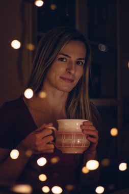 Güzel, küçük bir gülümseme ve pencereden dışarı bakan Avrupalı etnik kökenli genç bir kadın. Elinde sıcak çayla dolu büyük bir bardak tutan kız, stresten kurtulup özgürlük, huzur ve rahatlama duygusunun tadını çıkarıyor..