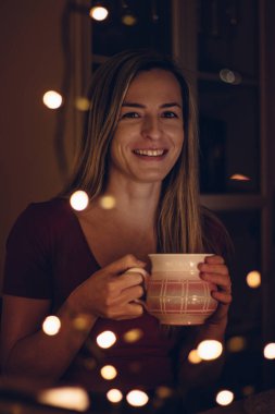 Kırmızı tişörtlü güzel bir mankeni çevreleyen sihirli sıcak ışık bir kupa sıcak çay tutuyor ve Noel 'in ilk pazar günü Noel ışıklarında gülümsüyor..