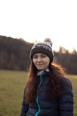 Gün batımında 20-24 yaş arası genç bir kızın hoş gülümsemesi. Kahverengi saçlı, güzel siyah beyaz şapkalı ve mavi ceketli bir kız. Köpeğini izliyor..