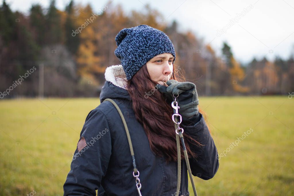 young dog breeder walk her dog. Detail on attention of a brunette who whistles disobedient dog. Woman aged 20-24 holds whistle and watches bitch. Candid portrait in authentic setting in grey moss tone.