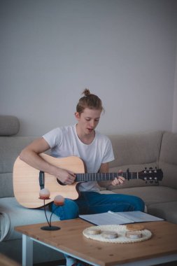 Sarı saçlı ve beyaz tişörtlü gerçek bir adam ışıklı bir odada koltukta oturup ahşap bir akustik gitarla akorları öğrenmeye çalışır. Notları okuyup pratiğe dökmek..