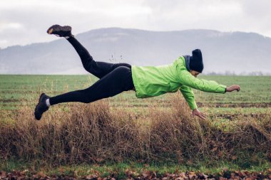 Yeşil kışlık ceketli atletik adam bir süpermen gibi sahaya atlıyor. Çimlerin üzerinde jimnastik figürü. Komik atlayış. Parkta genç bir adamla oynaşmak..