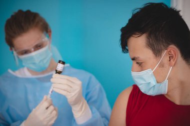 View of a patient in a red T-shirt waiting for vaccination and a doctor preparing a vaccine for a syringe. Vaccination center. Scary look of a young man. clipart