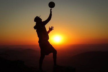 Hafif bir ceketi ve zirvesi olan bir sporcu bir kütüğün üzerinde duruyor ve gün batımında bir voleybol topuyla kafasının üstünde oynuyor. Spor dünyasının tadını çıkarıyorum..