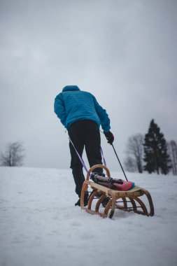 Mavi kış kıyafetleri içindeki sporcu, fotoğraf için aksesuarlı ahşap bir kızağı ve plastik bir snowboard 'u bayırın tepesine çekiyor. Kış ortamının ve baharda dağların samimi portresi.