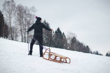 Mavi ceketli ve beyaz şapkalı genç adam denge için yürüyüş çubuklarını kullanarak tepeden aşağı koşuyor ve düşmeden daha iyi iniyor. Avrupa 'da kış dağı. Çocuklar yamaçlarda aylaklık ediyorlar..