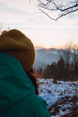 20-25 yaşlarındaki güzel genç bayan Çek Cumhuriyeti 'ndeki dağlarda güzel bir kış günbatımına bakıyor. - Günün sonunda. Sarı başlık ve yeşil ceket..
