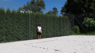 Beyaz tişörtlü ve siyah şortlu genç voleybol oyuncusu bir voleybol servisi gösteriyor. Sıçrama servisinin tanıtımı. Sahilde servis yapan genç bir atlet.