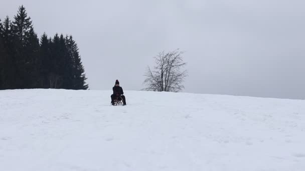 身穿黑色冬衣的村民骑着木制雪橇下山 他正试着用他的脚修正方向 冬日快乐23岁的男孩 回到他的童年 — 图库视频影像
