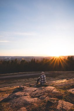 Yalnız gezgin Oslo yakınlarındaki kayalık tepede oturur ve ormanın ve fiyordun üzerindeki altın gün batımını seyreder..