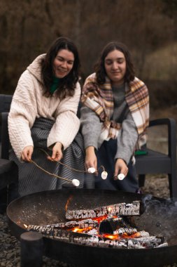 İki kadın kamp ateşinin önünde oturmuş şekerleme pişirirken düşünceli bir şekilde gülümsüyor.
