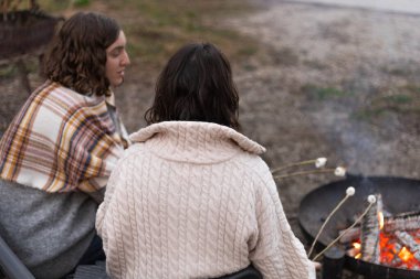 İki genç kadın kamp ateşinde marşmelov pişiriyor.