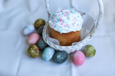 Süslü kumaşlı beyaz bir sepetin içinde Paskalya pastası, parlak boyalı yumurtalarla çevrili, bahar kutlamalarının sembolü.