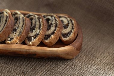 Çuval desenli uzun ahşap servis tabağında dilimlenmiş haşhaş tohumu rulosunun ılık tonlu fotoğrafı.