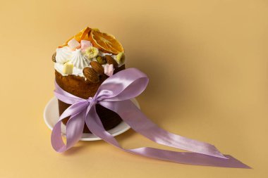 Badem, marşmelov ve portakal dilimleriyle süslenmiş paskalya pastası, pastel şeftali arka planında narin mor kurdeleyle bağlanmış.