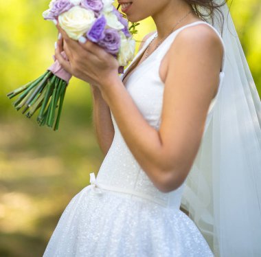 Değişken odak noktası, yeşil bir bahçenin arka planına karşı beyaz elbiseli bir gelinin elindeki buketin yakın plan görüntüsü. Açık havada düğün töreni.