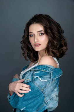 Yüzünde ciddi bir ifade olan esmer bir kadının yakın plan portresi beyaz iç çamaşırlı ve gri arka planda kot ceketli kameraya yan bakıyor.