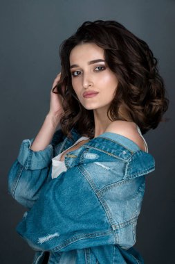 Yüzünde ciddi bir ifade olan esmer bir kadının yakın plan portresi beyaz iç çamaşırlı ve gri arka planda kot ceketli kameraya yan bakıyor.