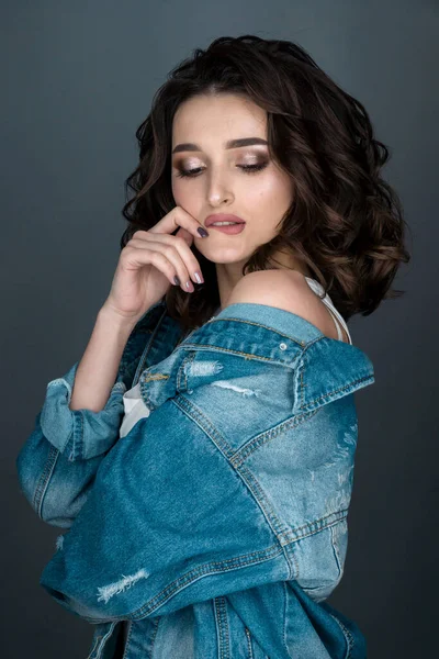 close-up portrait of a young sexy brunette who bit her lower lip and covered her eyes in pleasure, in white underwear and a denim jacket on a gray background
