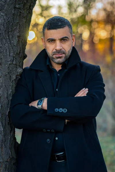 stylish man in black clothes stands in the park by the tree at sunset.