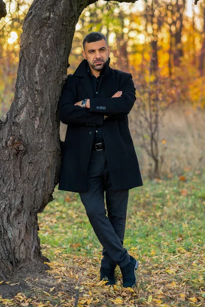 stylish man in black clothes stands in the park by the tree at sunset.