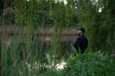 Siyah şapkalı genç sakallı adam nehrin kenarında duruyor, dönen bir çubukla balık tutuyor, yarım yamalak bir manzara, yemyeşil çimlerle çevrili.