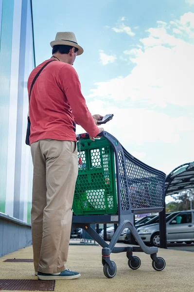Person walking holding mobile smart phone in hand during shopping. Cart on store car park background