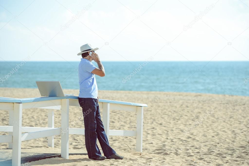 Busy male working on the laptop and talking on mobile. Man in hat speaking on smartphone over blue sky and ocean beach