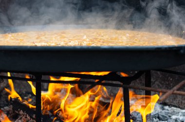 Authentic Spanish paella cooking over an open wood fire outdoors. Steam rising from the pan as rice simmers in broth. Concept of Mediterranean cuisine, traditional cooking, and rustic lifestyle.