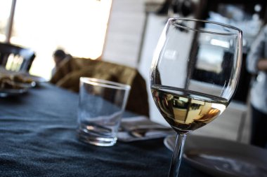 Yemek masasında beyaz şarapla dolu bir şarap kadehinin yakın plan fotoğrafı, bulanık bir arka plan ve rahat bir ev ışığı. Kutlama, öğle yemeği ya da gevşemiş sosyal toplantı kavramı.